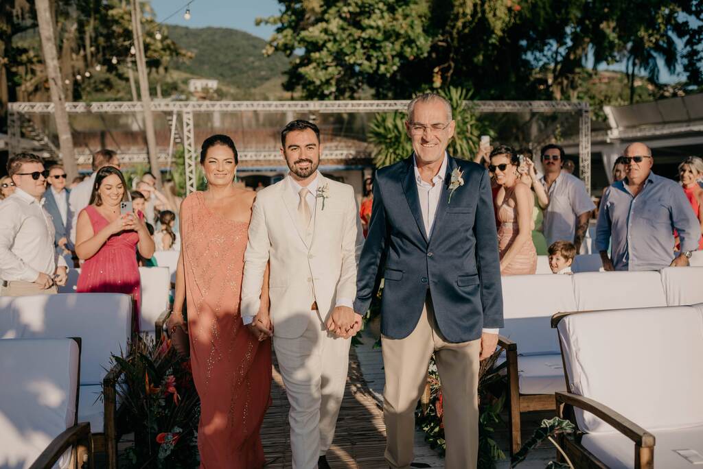 entrada do noivo com terno off white de mãos dadas com a mãe com vestido rosê longo com manga única e o pai com terno bege com terno azul acetinado