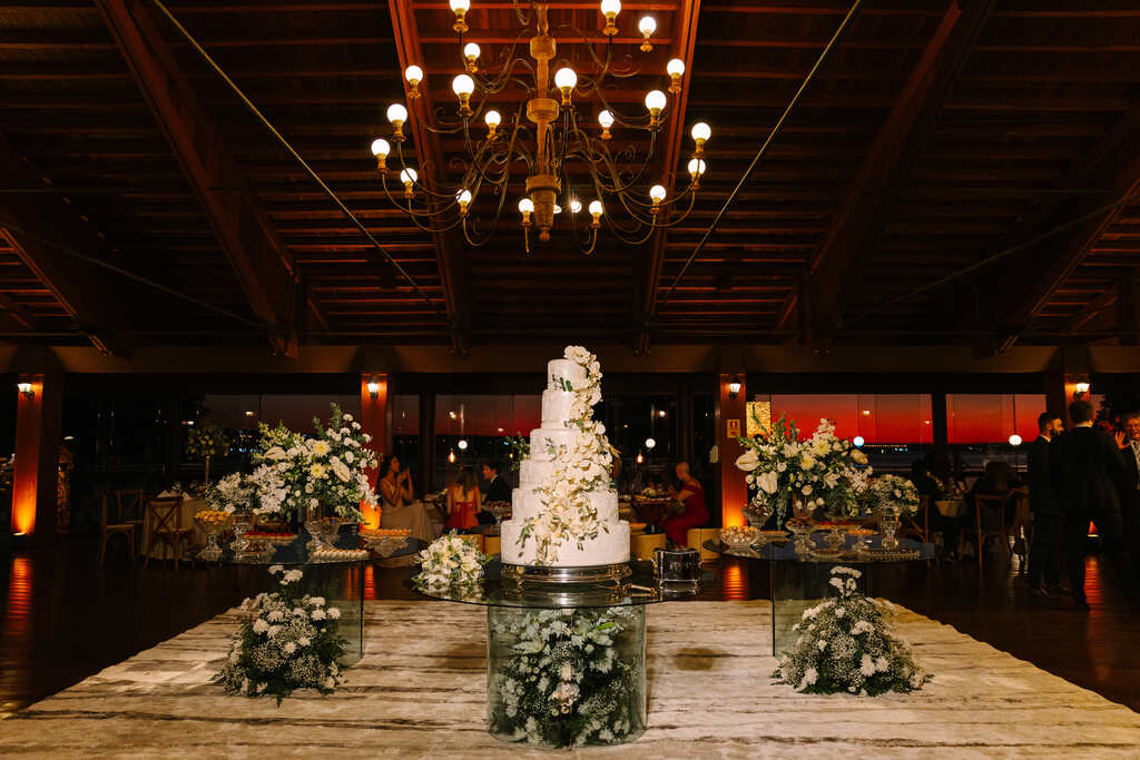 salão com mesas de vidor com arranjos de flores brancas e bolo de casamento com seis andares