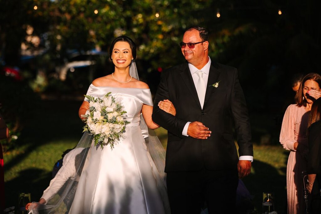 entrada da noiva com vestido minimalista com decote ombro a ombro segurando buquê branco ao lado do pai com terno preto