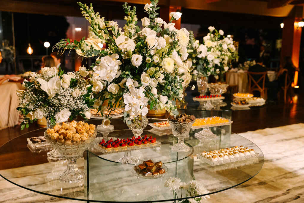 mesa redonda de vidro com vasos de flores brancas e rosas e bandejas com doces de casamento