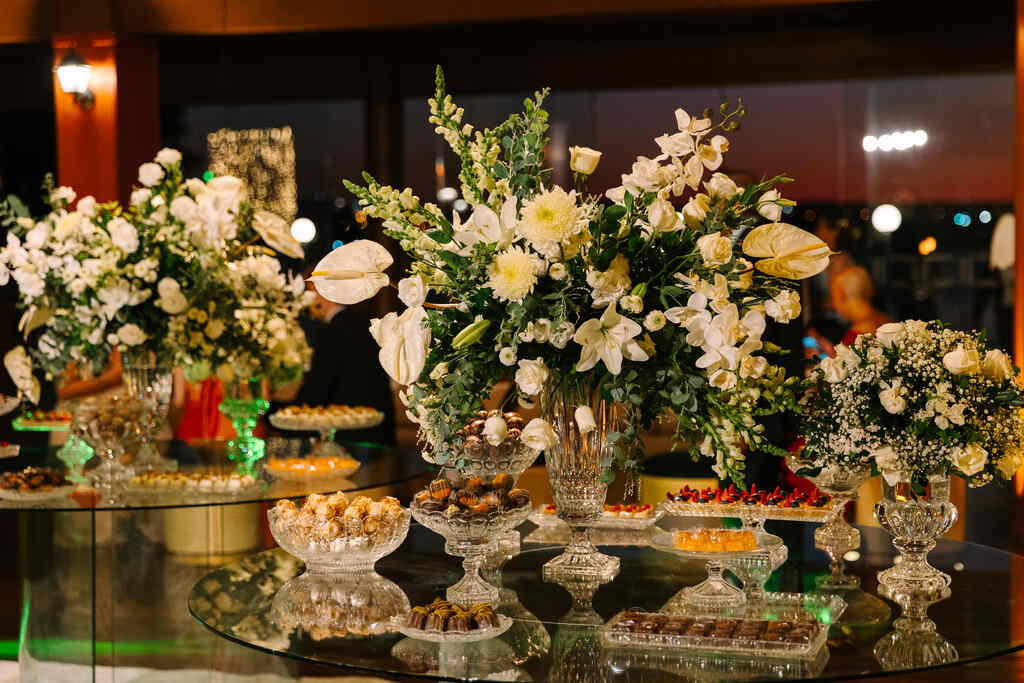 mesa redonda de vidro com vasos de flores brancas e rosas e bandejas com doces de casamento