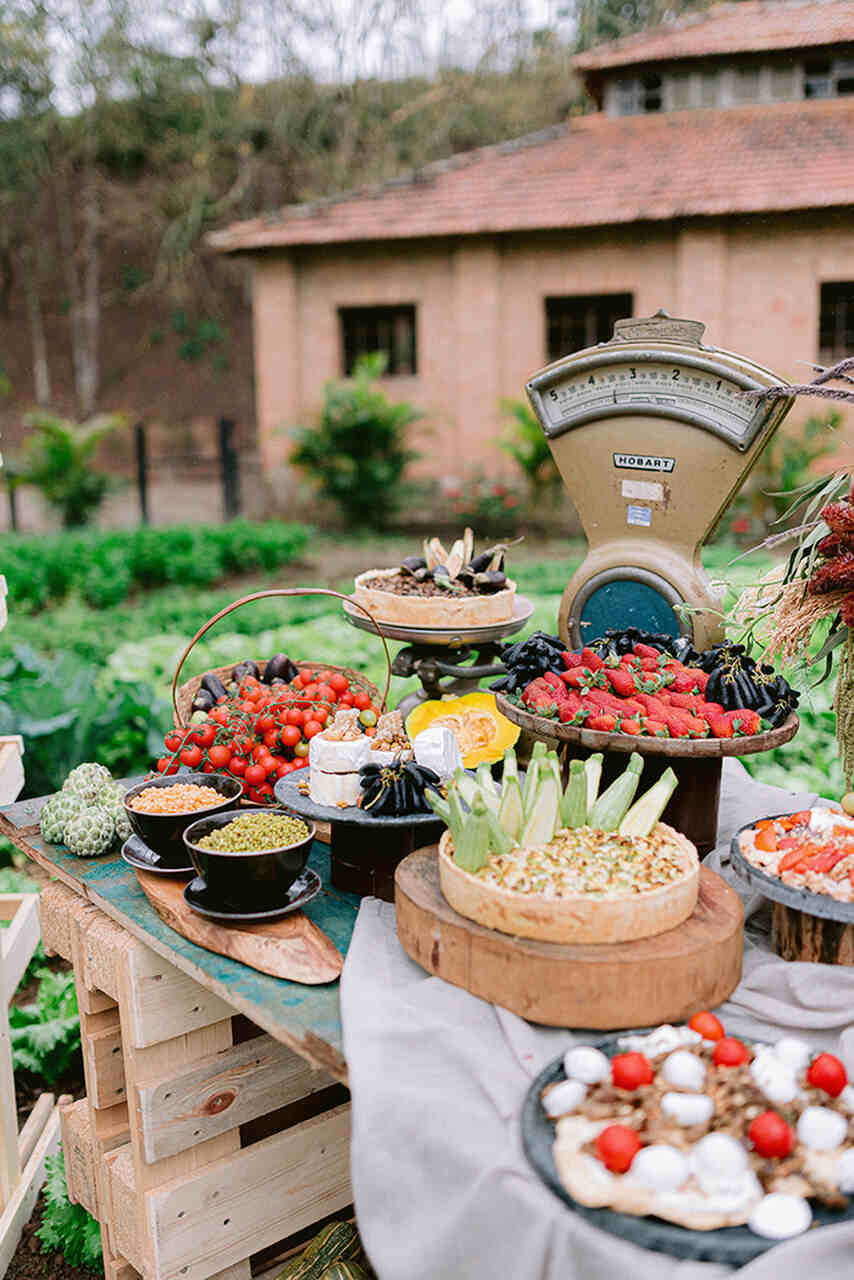 mesa com buffet para casamento no s´tio com balança antiga com uvas e morangos e ao lado tigelas com molhos cesta com tomates cereja e torta