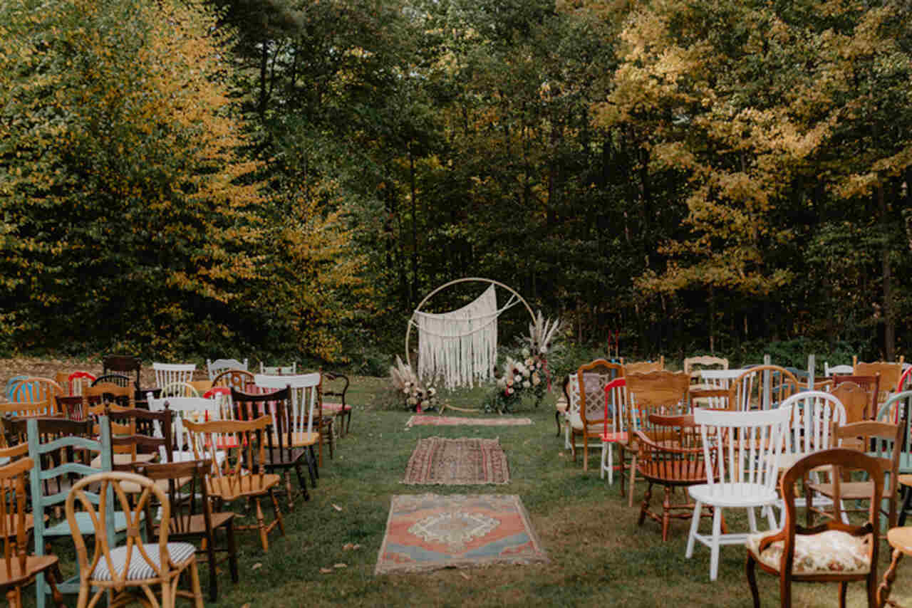 decoração para casameto no sítio com altar redondo com macramÊ e decorado com flores ao lado cadeiras de madeira em diversos estilos e cores e no caminho da noiva tapetes étnicos