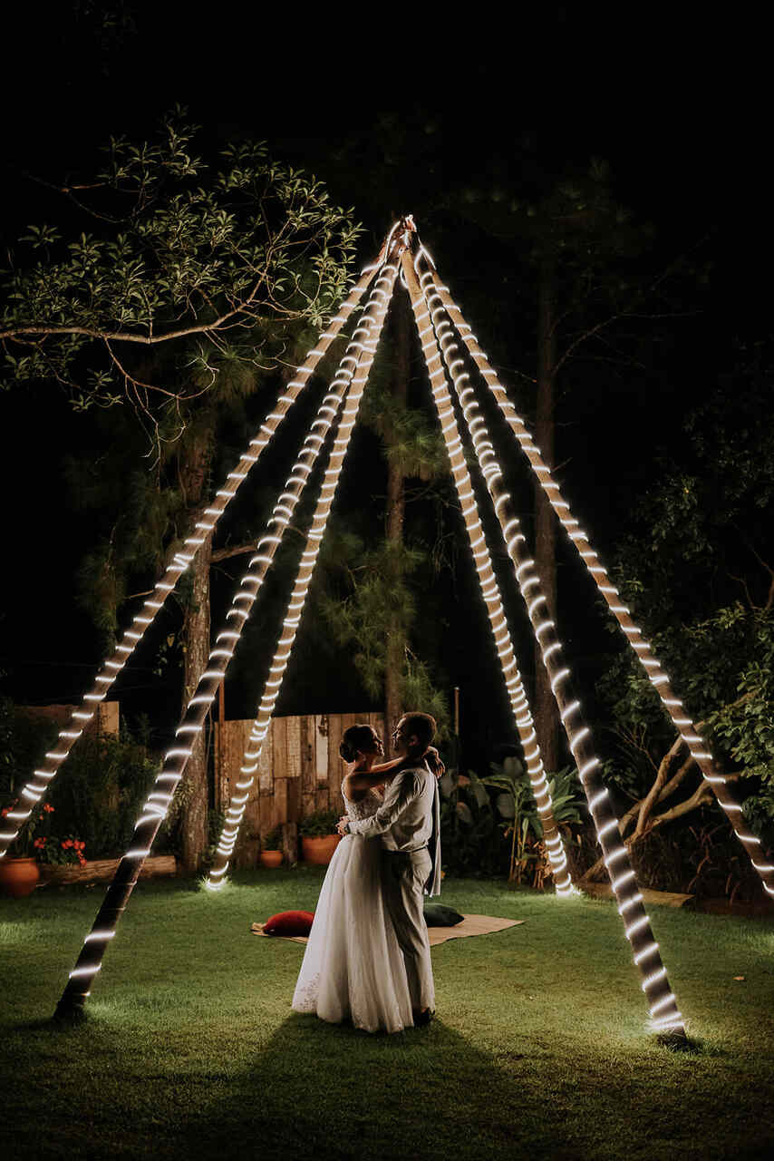 noivos abraçados embaixo de cabana de madeira decoradas com luzes à noite no sítio