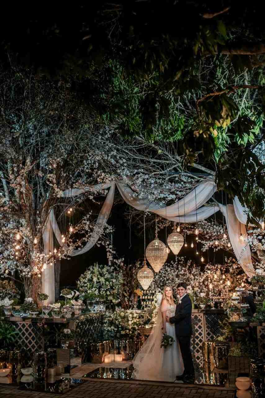 noivos na frente da mesa do bolo decorada com flores luminárias e palahas e tecidos pendurados nas árvore no jardim à noite