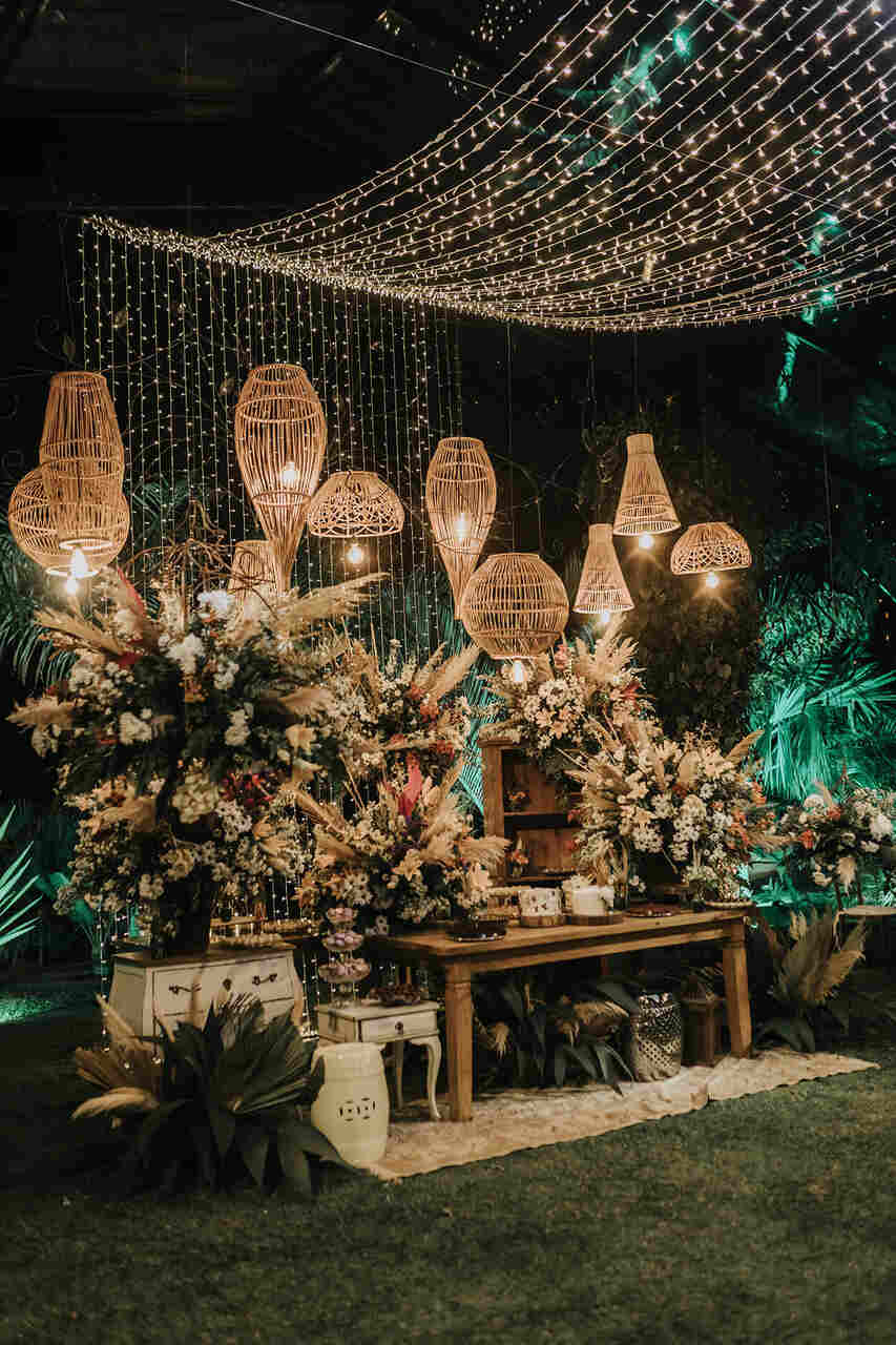 mesa do bolo com vasos de flores e luminárias de palha e varal de luzes por cima à noite