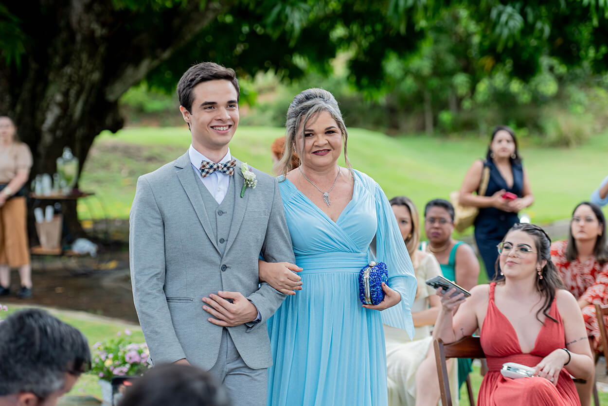 entrada do noivo com terno cinza com colete ao lado da mãe com vestido azul serenity