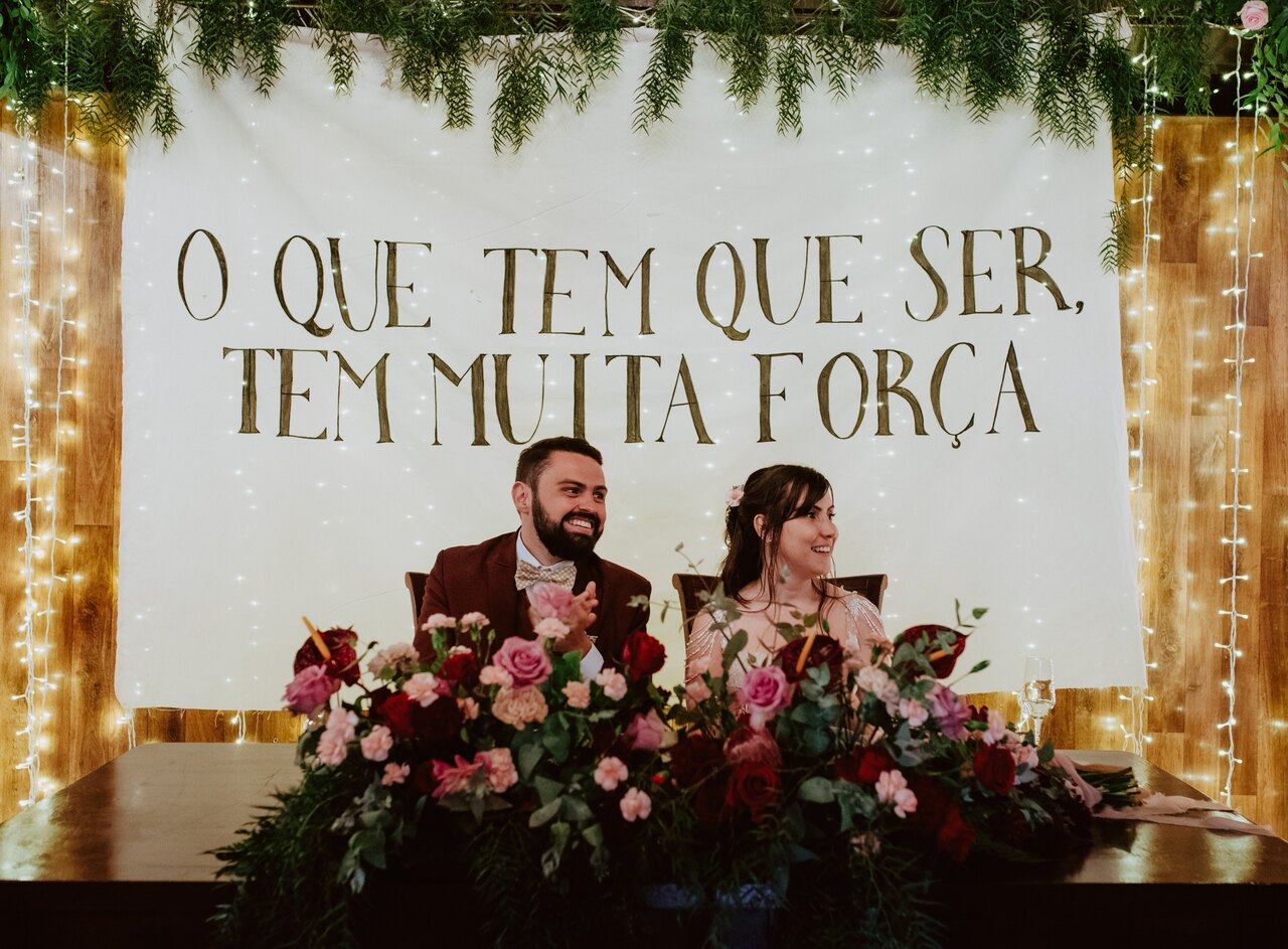 noivos sentados na mesa com flores vermelhas e rosas no centro e painel com tecido escrito o que tem que ser tem muita força
