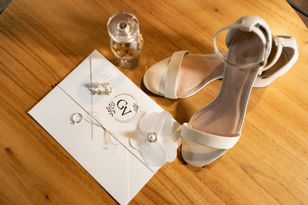 convite de casamento branco ao lado de vidro de perfume e sapato de noiva branco