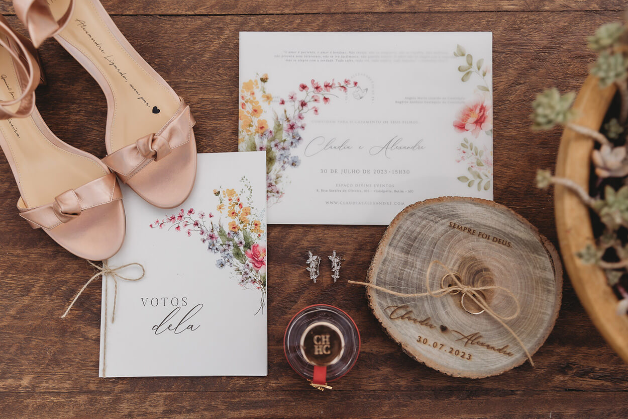 convite de casamento e caderno de votos branco com estampa de flores ao lado de sandália rosa brincos perfume e bolacha de madeira como porta alianças