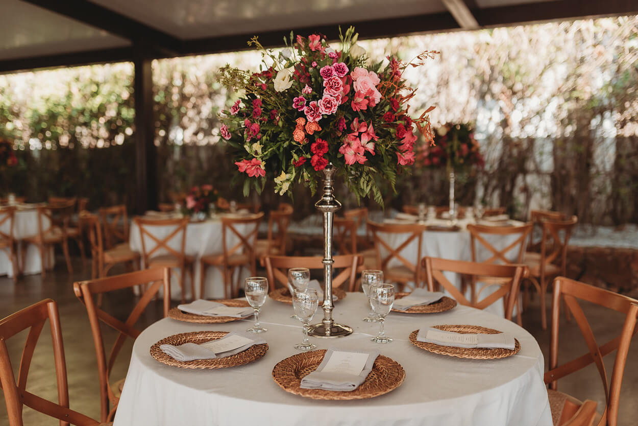 mesa redonda posta com toalha branca e sousplat de palha com flores rosas no centro da mesa