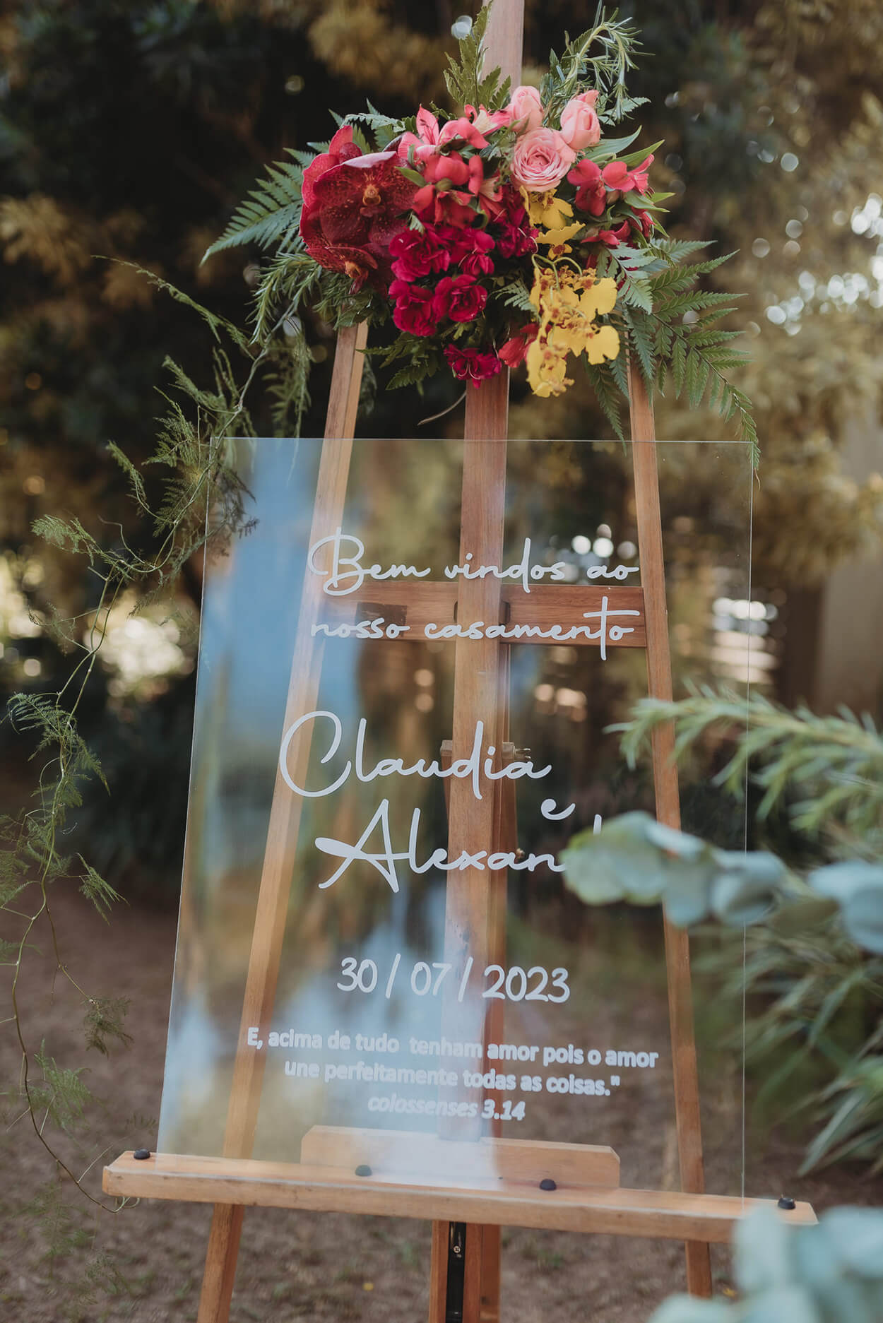 cavalete de madeira com quadro de acrílico transparente com o nome dos noivos e arrnjo com flores rosas e amarelas acima
