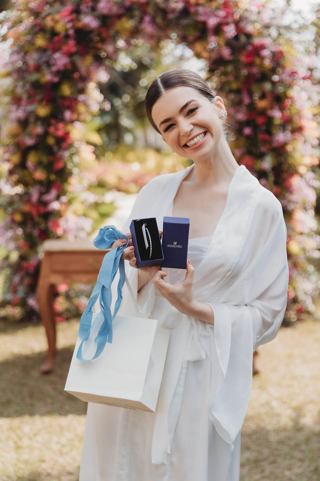 noiva com robe branco ao ar livre sorrindo enquanto mostra caixa de presente com pulseira