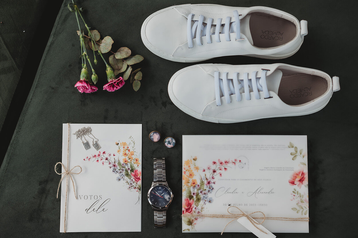 tênis masculino branco ao ado de flores rosas relógio prateado e convite de casamento e caderno de votos branco com estampa de flores 
