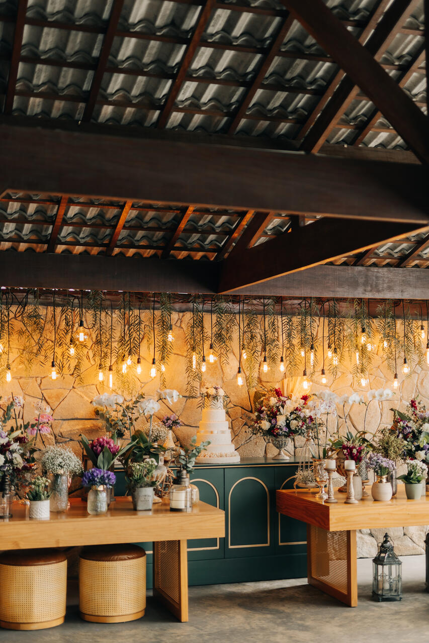 salão coberto com mesa verde com bolo de casamento e vários vasos com flores brancas rosas e lilás