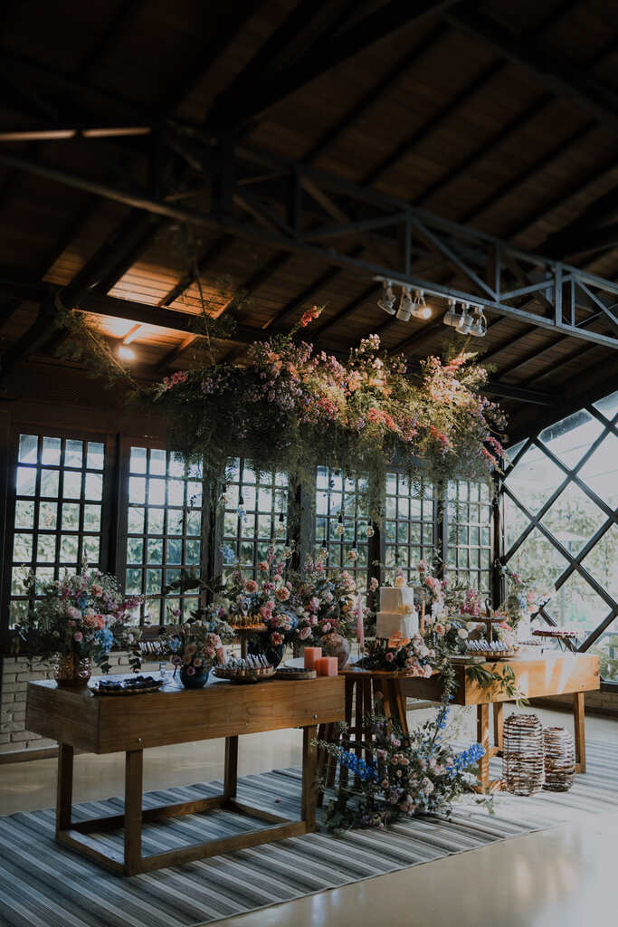 mesas de madeira com arranjos com flores rosas e azuis e bolo de casamento branco com dois andares no salão rústico
