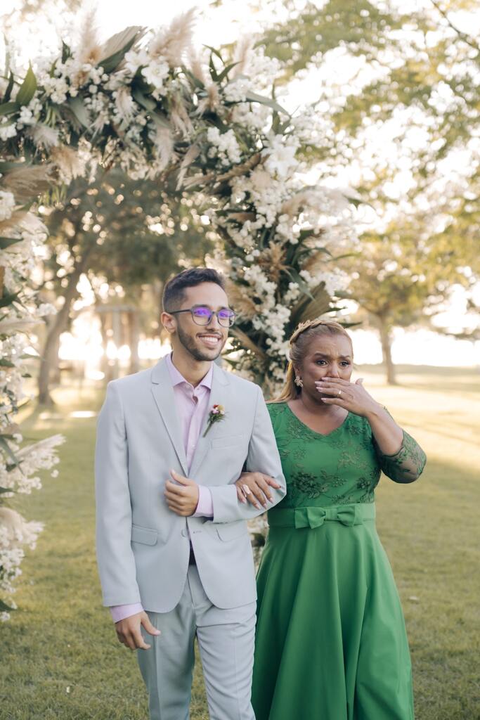 entrada do noivo com terno cinza e gravata rosa oa lado da mãe com vestido verde