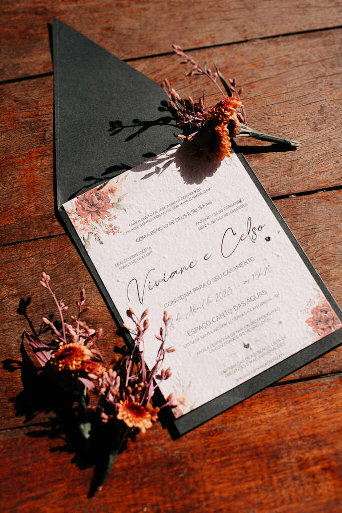 convite de casamento branco com estampa floral sobre envelope verde ao lado de flores secas em cima da mesa
