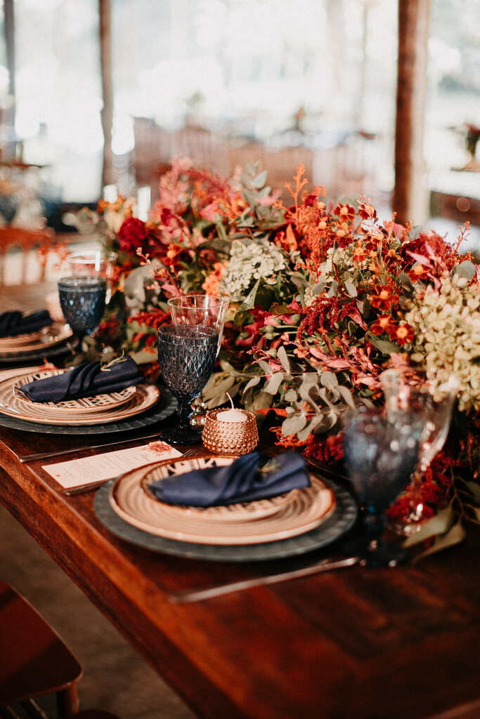 mesa posta de madeira com sousplat taça e guardanapos azuis e flores vermelhas e laranjas no centro