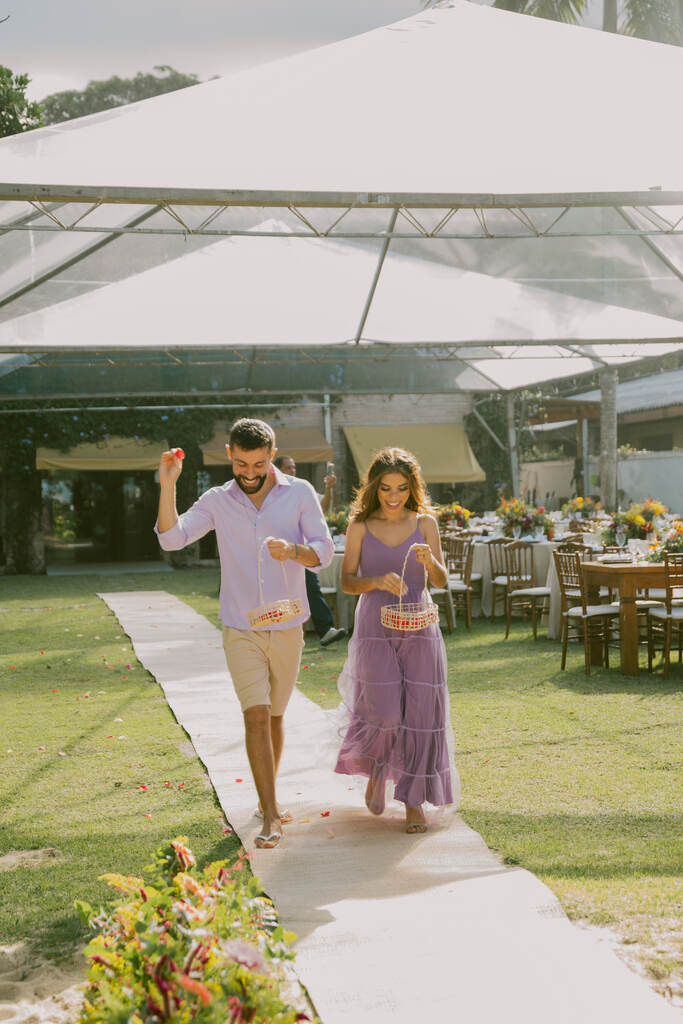 homem com bermuda jogando flores e mulher com vestido lilás segurando cesta rústica com flores