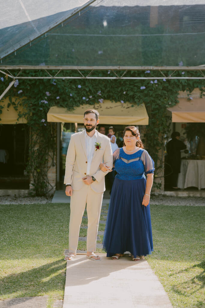 entrada do noivo com terno bege ao lado da mãe com vestido azul escuro