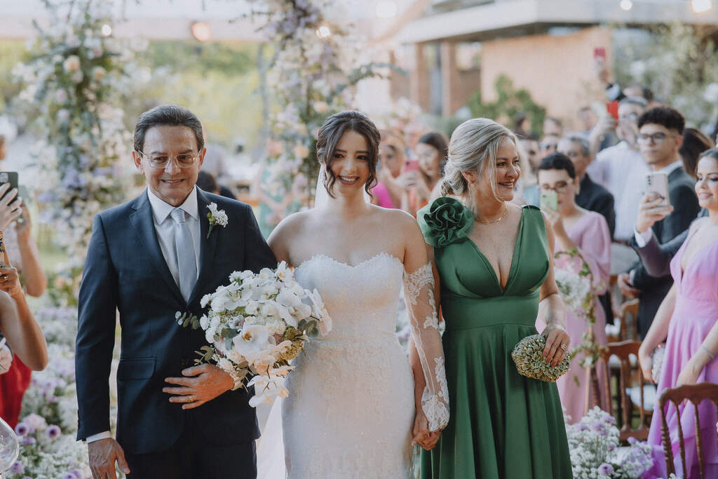 entrada da noiva segurando buquê branco ao lado do pai com terno azul escuro e mãe com vestido verde 