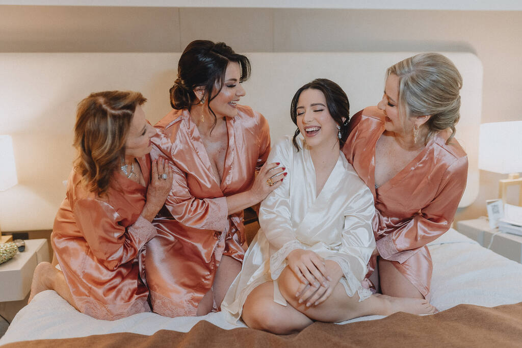 noiva com robe branco sentada na cama ao lado de três mulheres com robes rosas