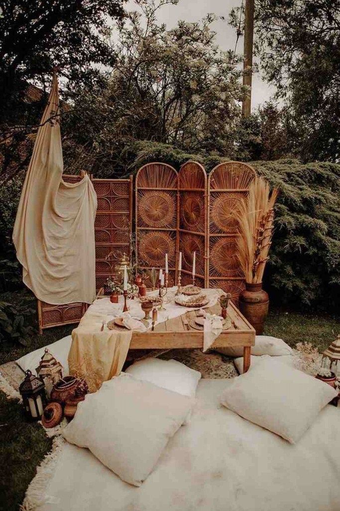 lounge para casamento no sítio com estilo boho com biombo de palha com tecido bege por cima mesa de centro de madeira decorada com velas e almofadas brancas no chão