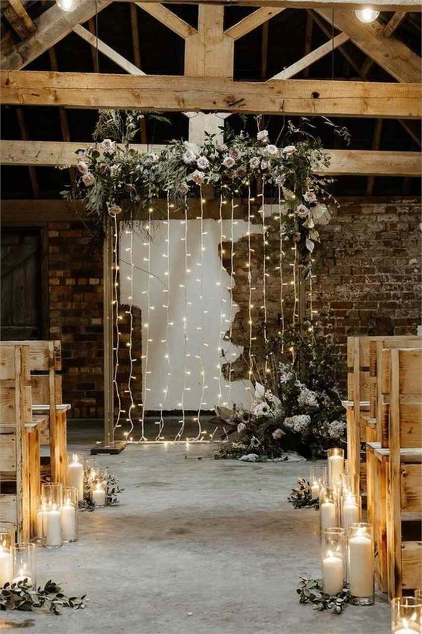 Altar com varala de luzinhas e flores penduradas no teto dentro de salão rústico com parede de tijolos e ao fundo bancos de madeira e velas no chão