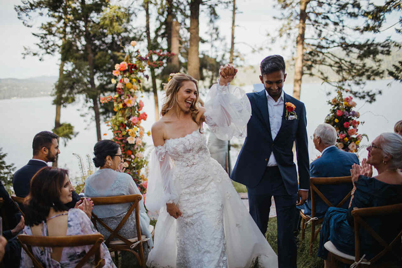 Mulher loira com vestido de noiva com manga de mãos dadas com o noivo comemorando o casamento no sítio com vista para o lago ao fundo convidados sentados em cadeiras de madeira aplaudindo 