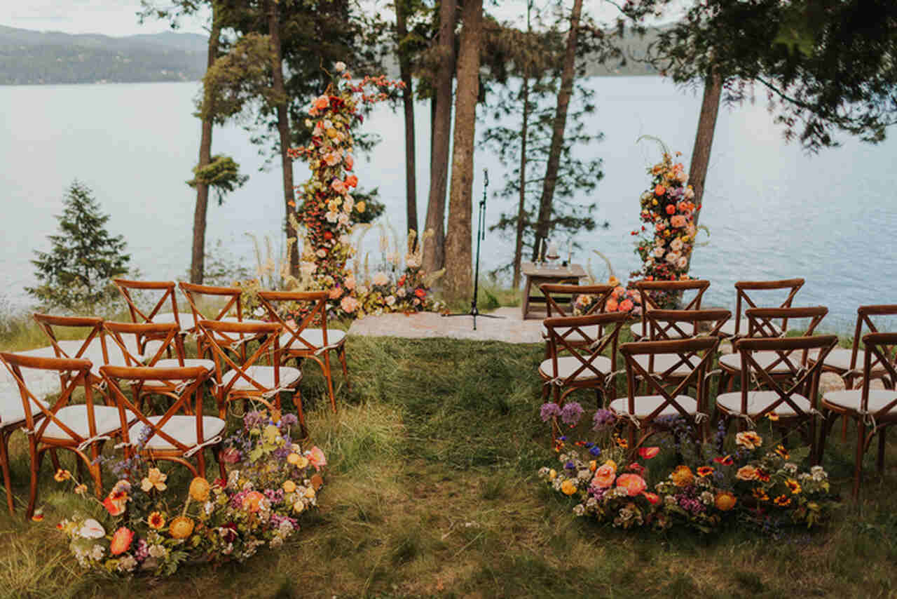 cadeiras de madeira decorada com arranjos de flores amarelas, lilás brancas e laranjas e altar desconstruído com flores coloridas debaixo da árvore com vista para o lago