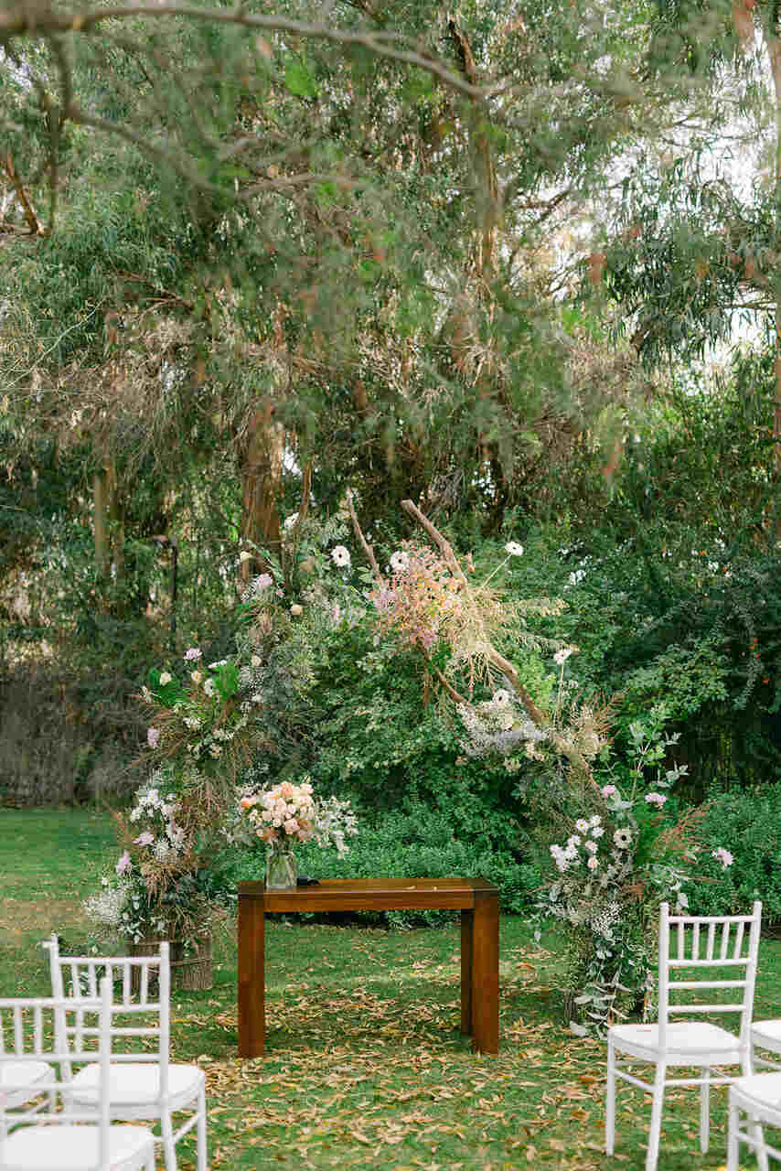 mesa de madeira simples e altar desconstruido com flores brancas e laranjas e ao lado cadeiras brancas estilo tiffany