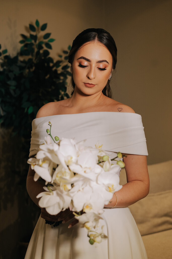 noiva com mquiagem simples e vestido de noiva com decote ombro a ombro segurando buquê com orquídeas brancas