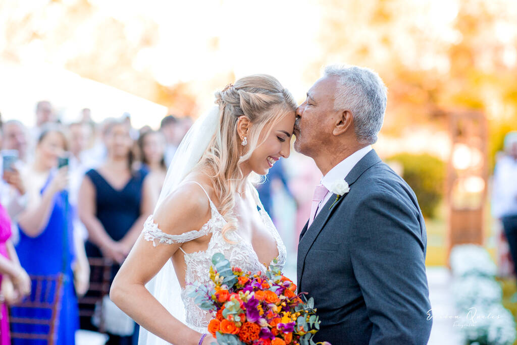 pai da noiva com terno cinza escuro beijando a testa da noiva segurando buquÊ com flores roxas laranjas e amarelas