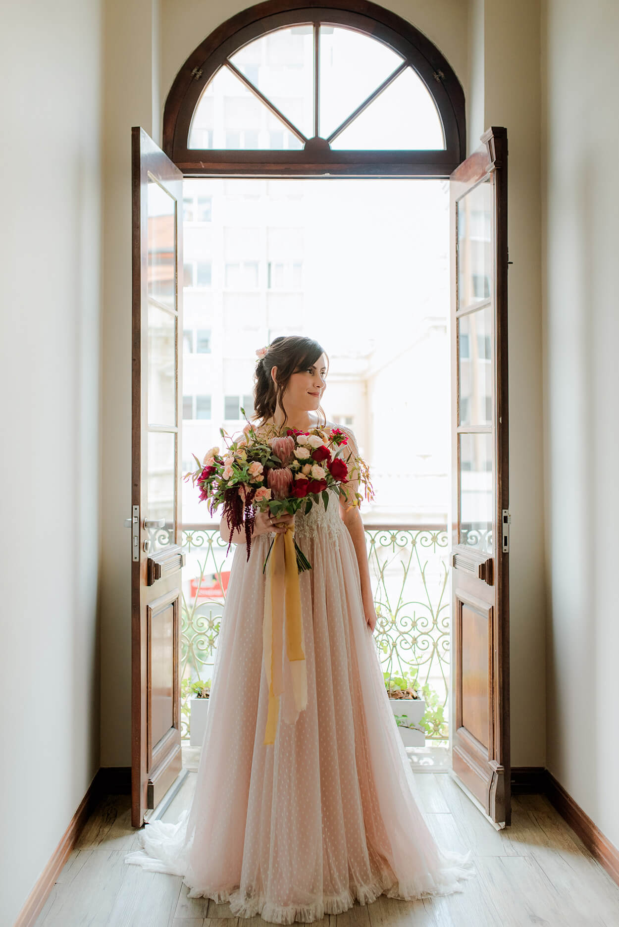 noiva na frente de porta aberta segurando buquê grande com flores vermelha brancas e proteas