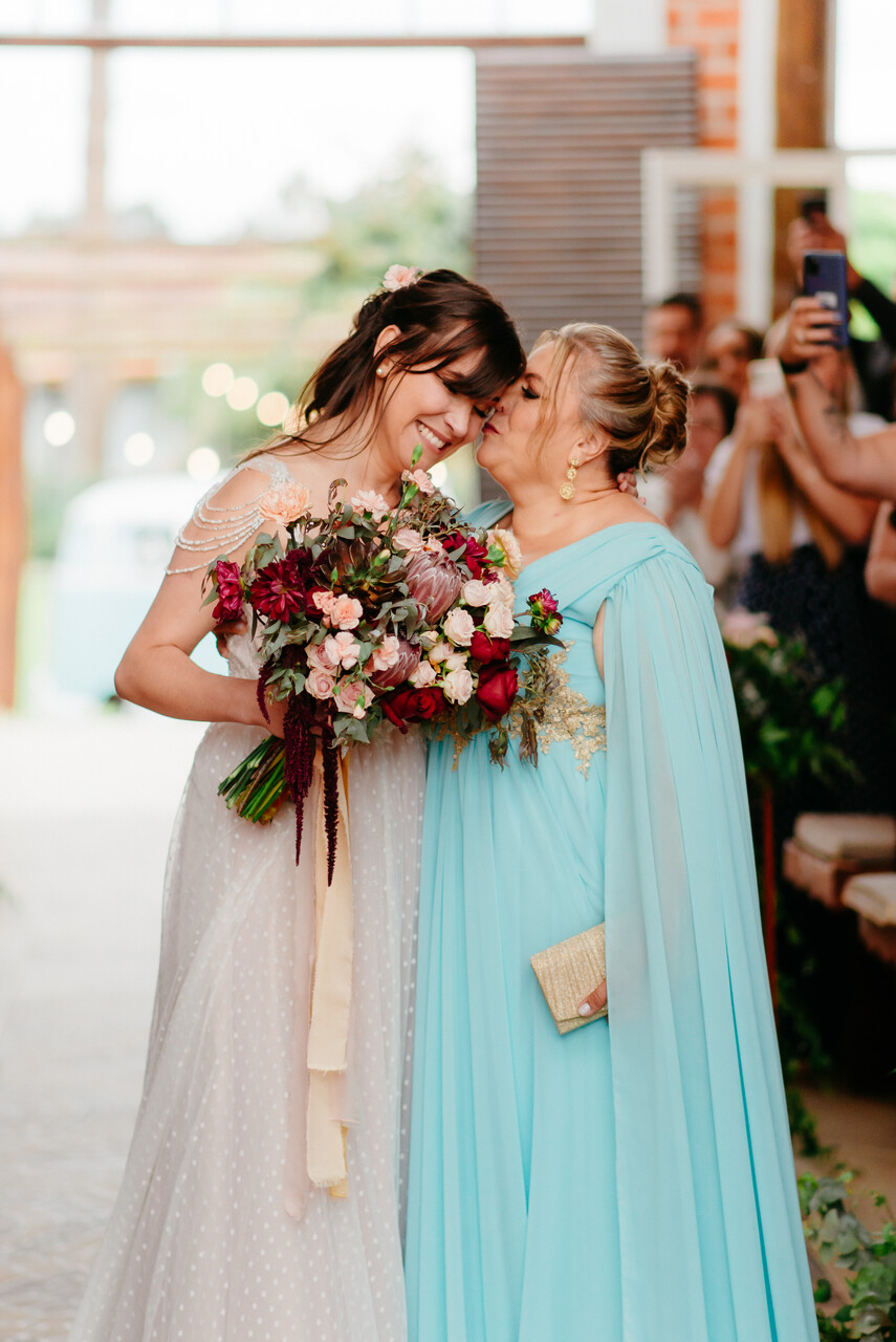 entrada da noiva segurando buquê enquanto recebe beijo do rosto da mãe vestindo vestido azul serenity com capa