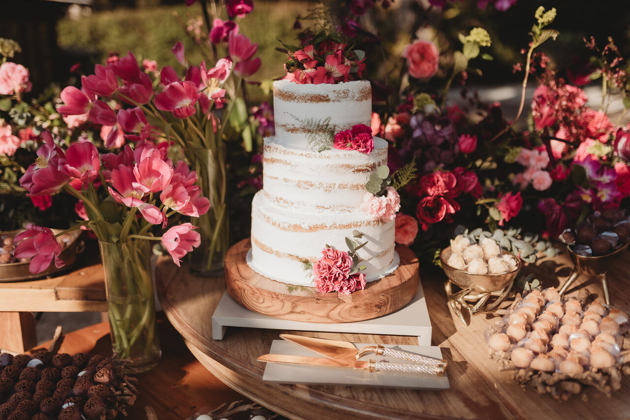 mesas de madeira com bolo de casamento semi espatulado com tres andares e arranjos de flores cor de rosa ao ar livre