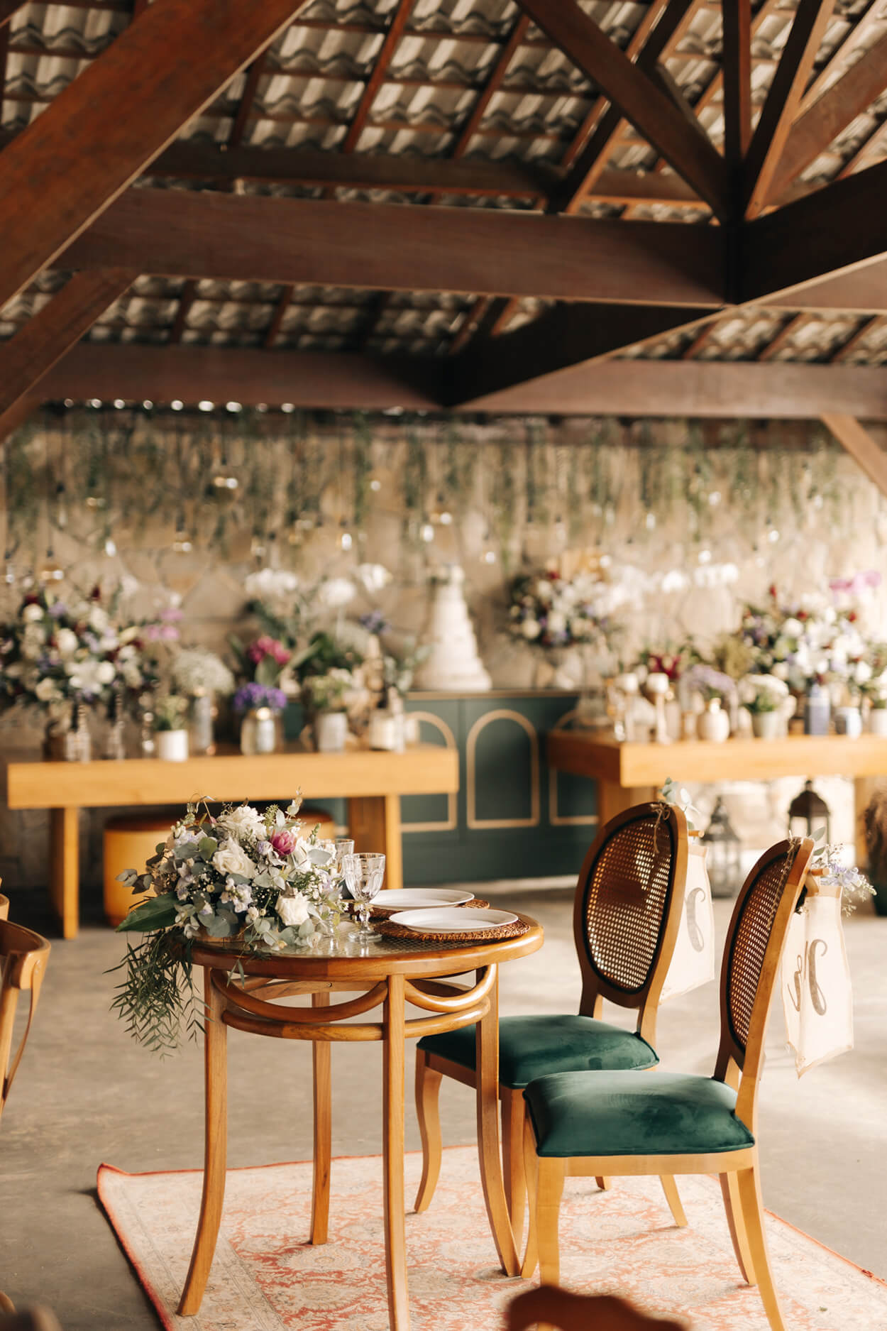 salão coberto com mesa redonda ppara os noivos com flores e ao fundo mesa verde com bolo de casamento e vários vasos com flores brancas rosas e lilás