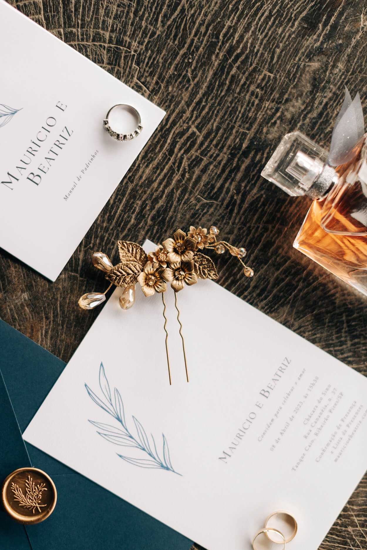 convite de casamento branco minimalista com envelope azul e lacre de cera dourado ao lado de perfume grinalda e brincos dourados