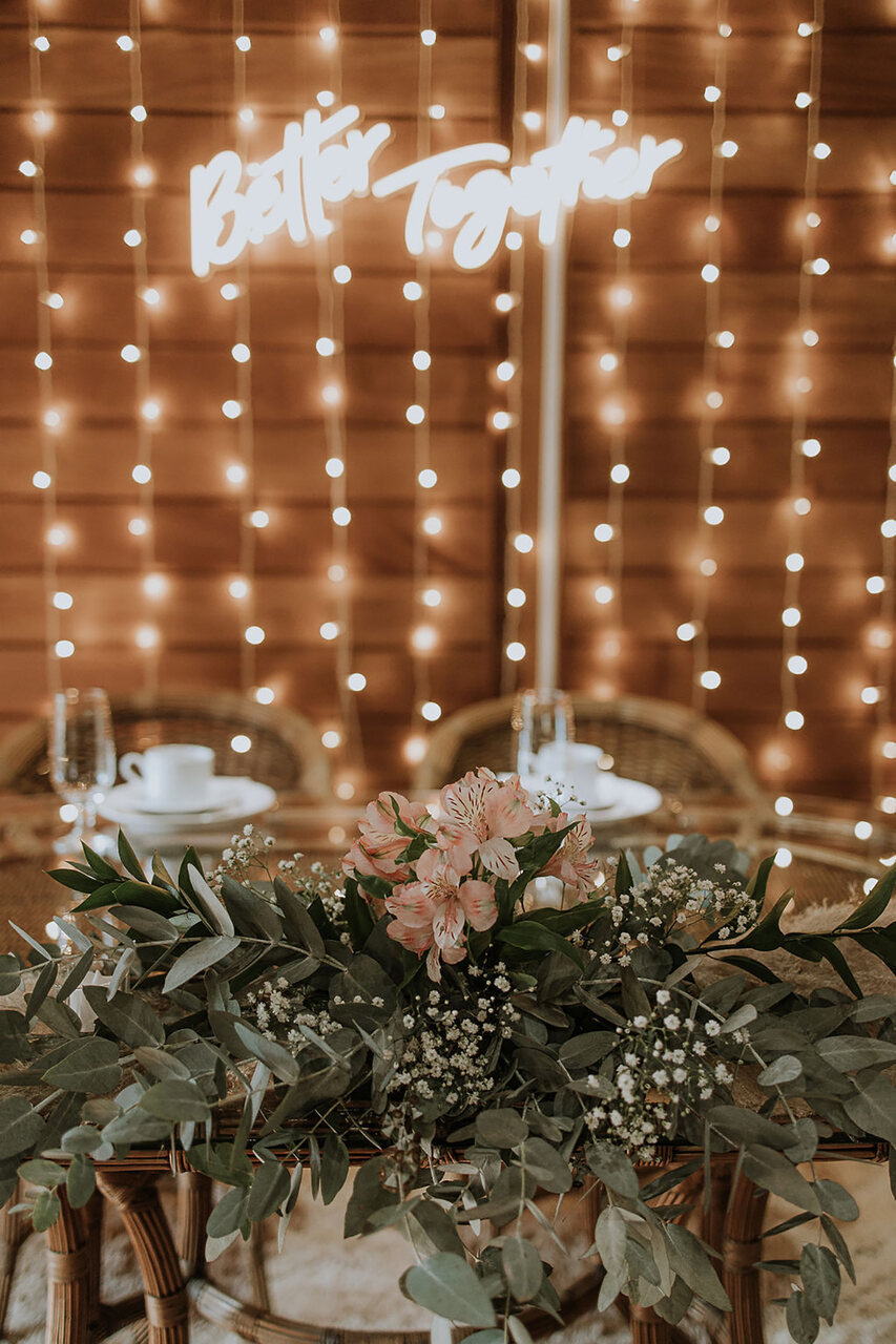 mesa com cadeiras de madeira com flores e atrás parede decorada com letreiro neon e varal de luzinhas