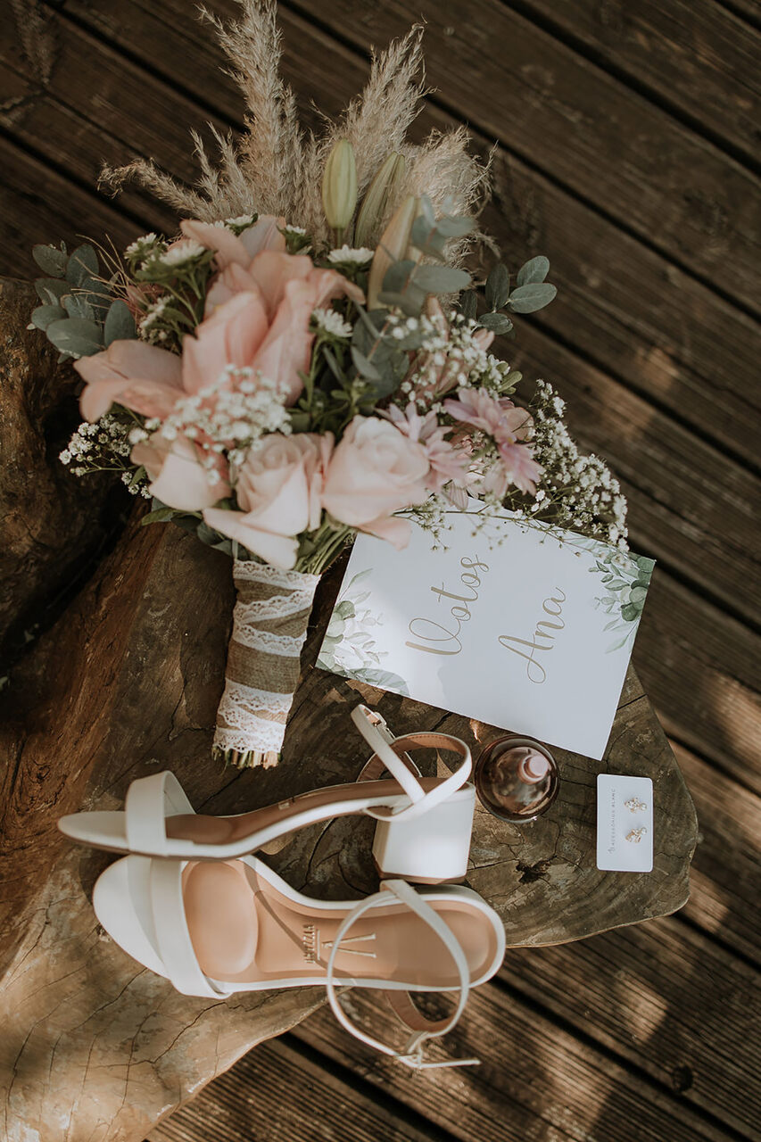 buquê com flores rosas e capim dos pampas e sandálias brancas ao lado do convite branco com folhagens