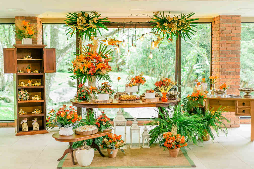 salão decorado com mesas de madeira trio de bolos de casamento e vários arranjos de flores laranjas e amarelas