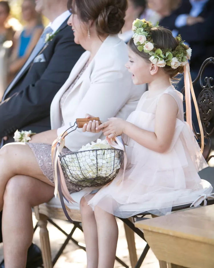 florista de casamento sentada observando a cerimônia segurando cesto de metal com pétalas brancas