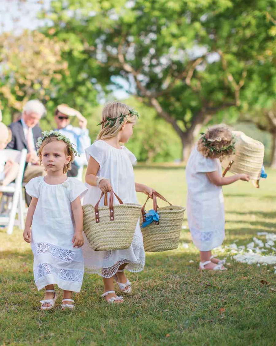três floristas de casamento com vestido branco, coroa de flores, sandalia e carregando bolsas de palha