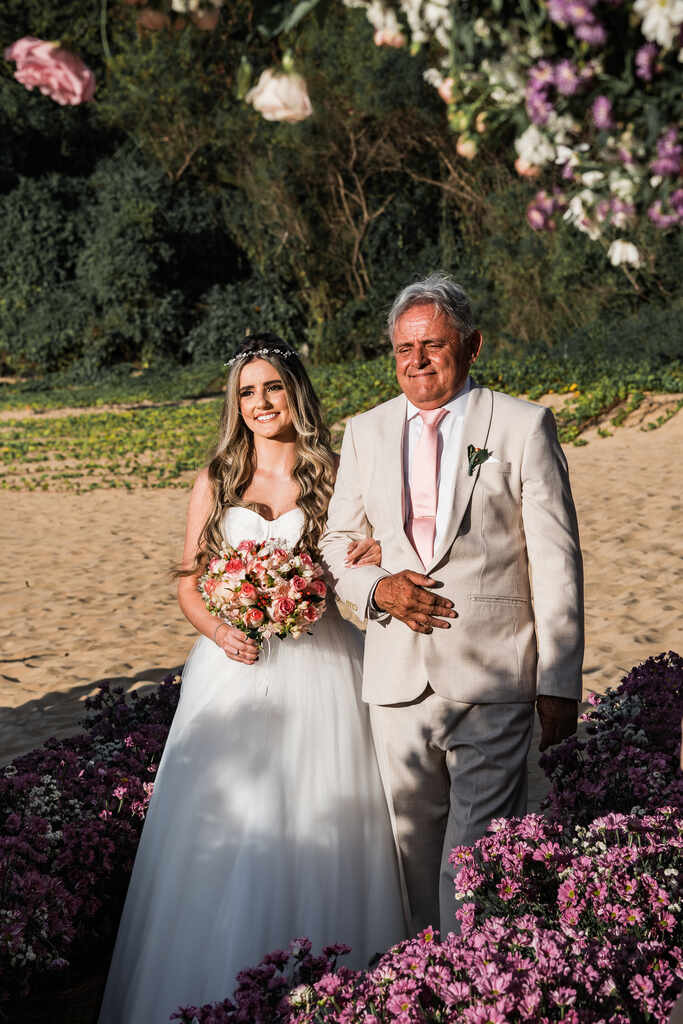 entrada da noiva segurando buquê redondo com flore sbrancas e rosas ao lado do pai com terno off white com gravat rosa clara na praia