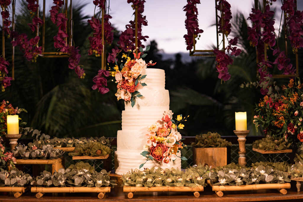 bolo de casamento espatulado com cinco andares com topo com flores rosas e laranjas e ao lado bandejas de madeira com doces de casamento e vasos com flores