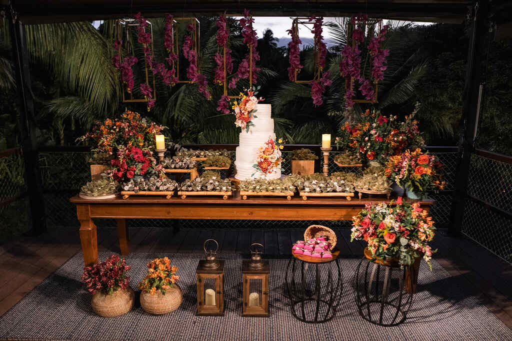 mesa de madeira com bolo de casamento com cinco andares com topo com flores rosas e laranjas e ao lado bandejas de madeira com doces de casamento e vasos com flores