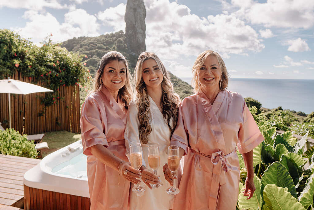 noiva com robe branco brindando com duas mulheres vestindo robe cor de rosa