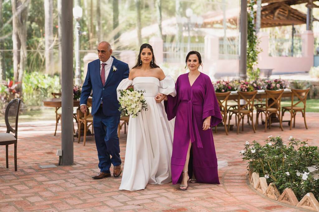 entrada da noiva segurando buquê com flores brancas de mãos dadas com o pai com terno azul escuro e gravata vermelha e mãe com vestido roxo