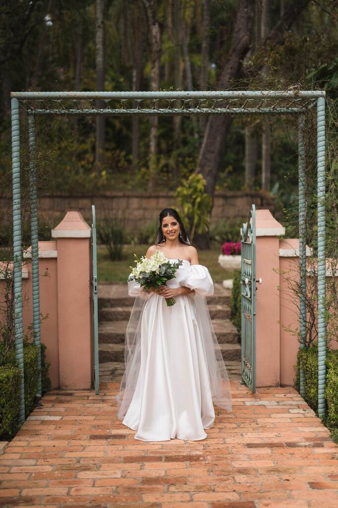 noiva com vestido minimalista com manga bufante segurando buquÊ redondo com flores brancas na frente da porta de fazenda antiga